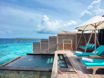 Luxuriöse Überwasservilla mit Holzdeck, blauem Pool, Liegen und türkisfarbenem Meer im Hintergrund.