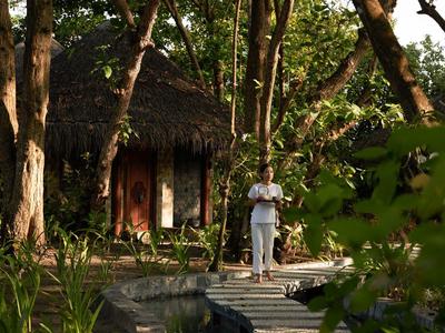 Ein Mann steht auf einem Holzsteg zwischen Bäumen vor einer Hütte mit Strohdach im Wald.