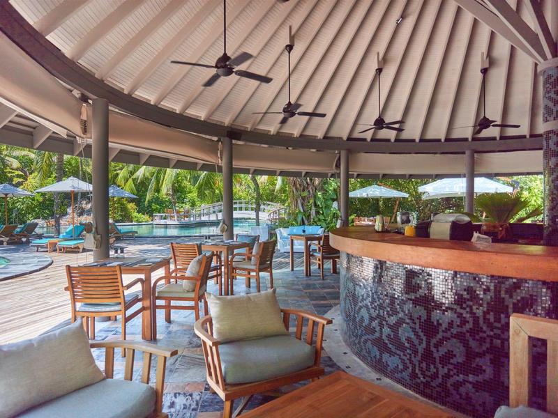 Offener Poolbarbereich mit Holzmöbeln, Deckenventilatoren und Blick auf einen Pool mit Liegestühlen.