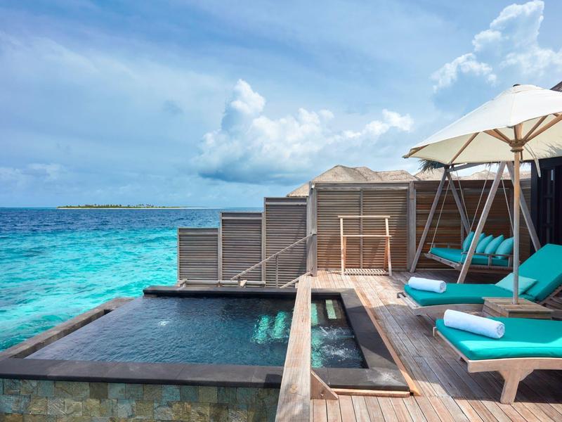 Luxuriöse Überwasservilla mit Holzdeck, blauem Pool, Liegen und türkisfarbenem Meer im Hintergrund.