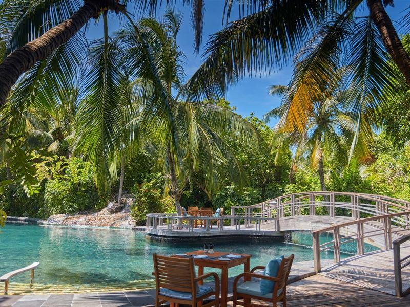 Ein tropischer Pool mit Holzbrücke, Palmen, blauem Himmel und Sitzbereich mit Tischen und Stühlen.
