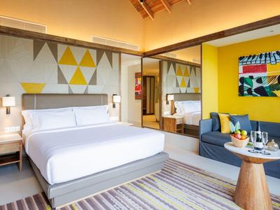 Helles Hotelzimmer mit großem Bett, gelber Wand, Sofa, modernem Kunstbild und Teppichboden.