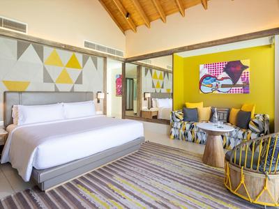 Helles Zimmer mit Holzbalkendecke, großem Bett, dekorativen Kissen, bunten Wänden und Teppichboden.