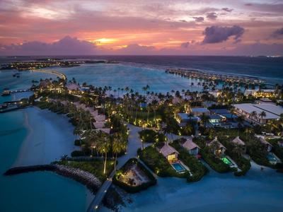 Insel mit vielen Palmen, Häusern und blauem Wasser unter rosa-orangefarbenem Himmel bei Sonnenuntergang.