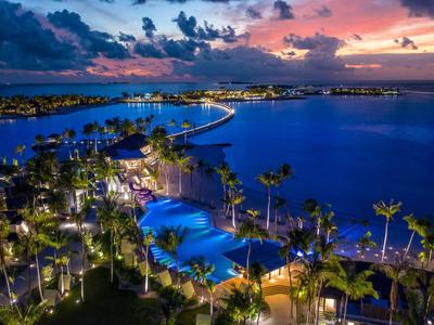 Blick auf beleuchtete Poolanlage und Palmen am Meer bei Sonnenuntergang mit farbigem Himmel.