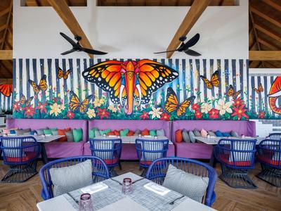 Buntes Schmetterlings-Wandbild in hellem Restaurant mit blauen Sesseln und Holzdachstuhl.