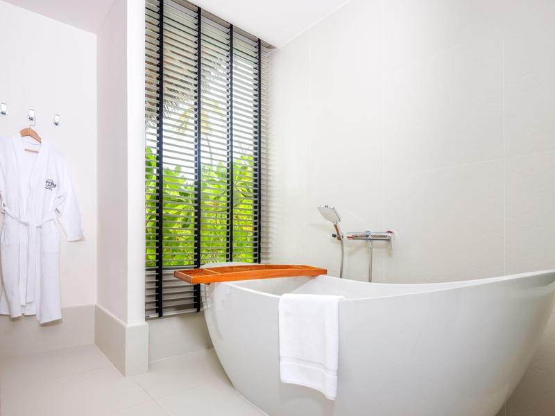 Helle Badezimmer mit freistehender weißer Badewanne, Handtuch, Holzablage und Fenster mit schwarzen Lamellen.