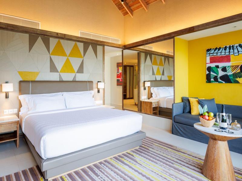 Helles Hotelzimmer mit großem Bett, gelber Wand, Sofa, modernem Kunstbild und Teppichboden.