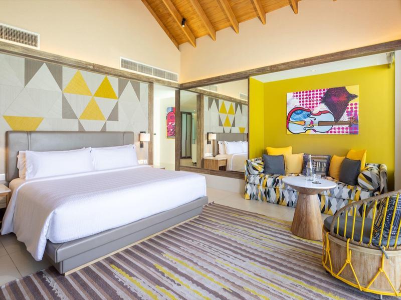 Helles Zimmer mit Holzbalkendecke, großem Bett, dekorativen Kissen, bunten Wänden und Teppichboden.