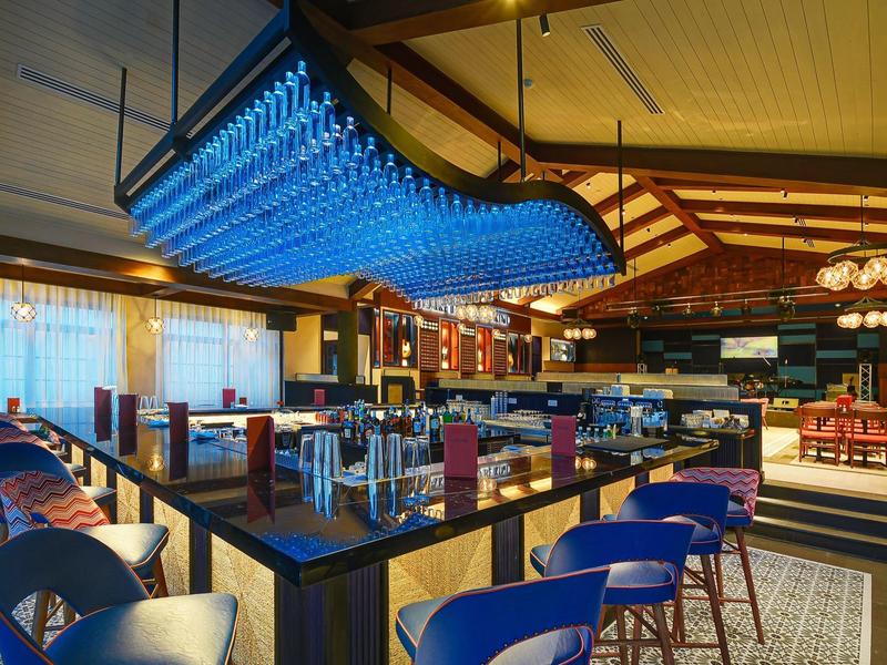 Moderner Barbereich mit blauen Hockern, Holztischen und einer markanten blauen LED-Deckenleuchte.