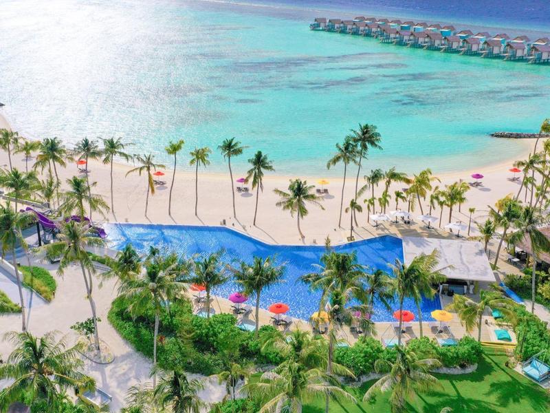 Türkisfarbenes Meer, weißer Sandstrand mit Palmen und großer blauer Poolanlage im Vordergrund.