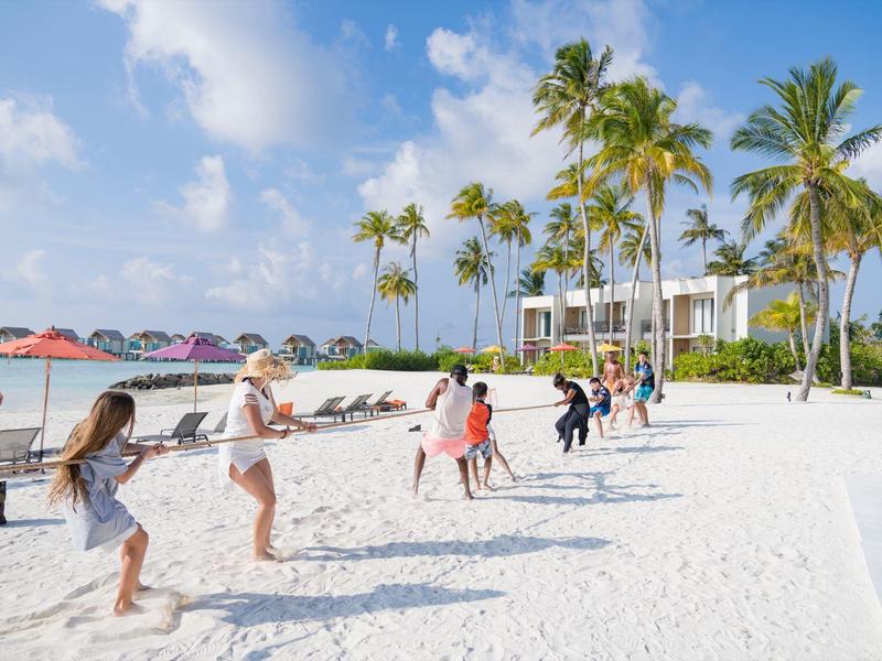 Kinder spielen Tauziehen am weißen Sandstrand unter Palmen bei sonnigem Wetter.