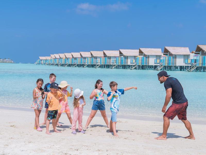 Kinder spielen am weißen Sandstrand nahe türkisfarbenem Wasser mit über dem Wasser gebauten Bungalows.