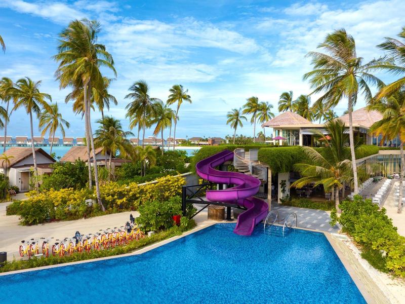 Großer blauer Pool mit lila Rutsche, Palmen und Strand unter blauem Himmel und weißer Wolken.