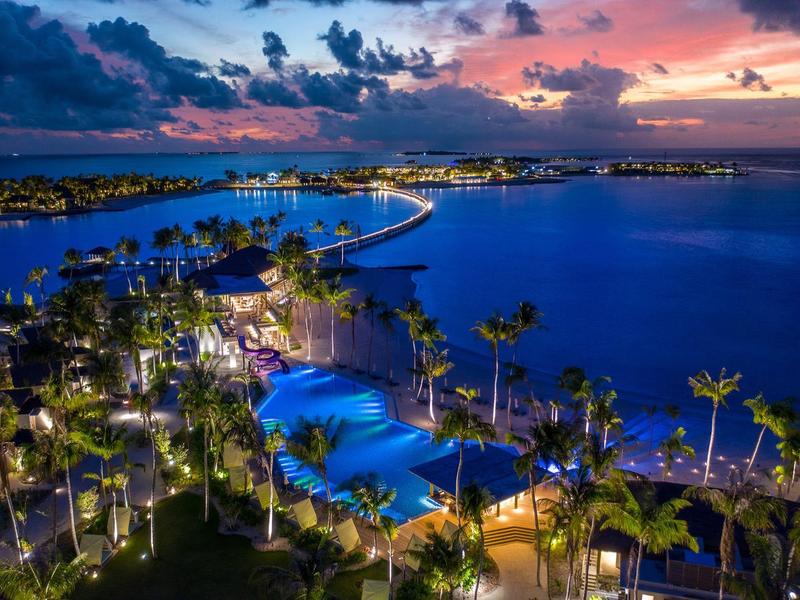 Blick auf beleuchtete Poolanlage und Palmen am Meer bei Sonnenuntergang mit farbigem Himmel.