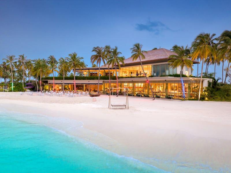 Strand mit weißem Sand, türkisfarbenem Wasser, Palmen und einem beleuchteten Restaurant bei Abenddämmerung.