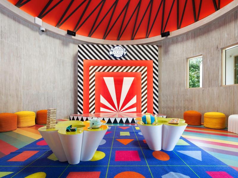 Bunter Kinderspielraum mit rundem roten Dach und Teppich, kleinen weißen Tischen und Sitzkissen.