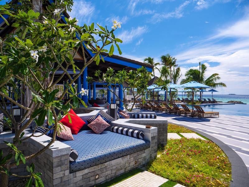 Zewnętrzny salon z tapicerowaną ławką, poduszkami, parasolami i leżakami w tropikalnej pogodzie.