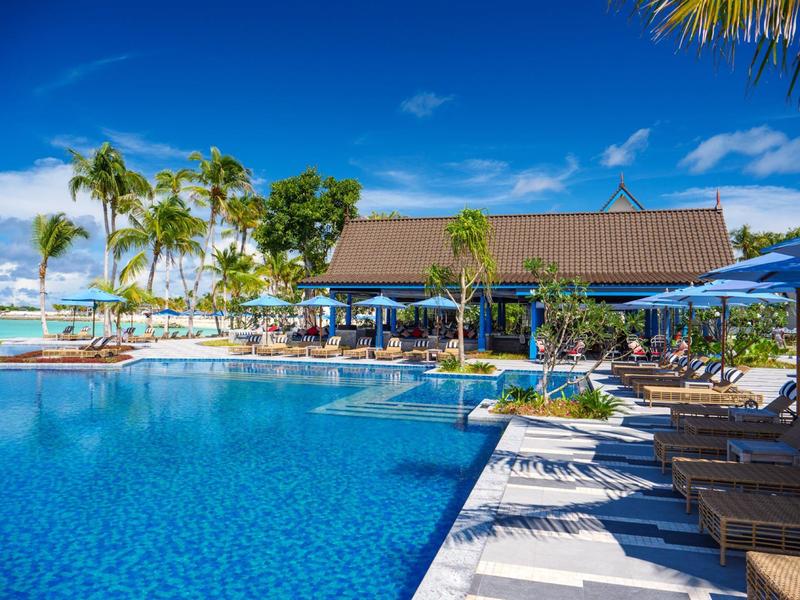 Duży basen z leżakami i palmami przed hotelem w słoneczny dzień.