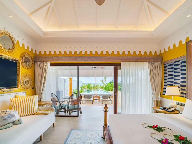 Jasny pokój hotelowy z żółtymi ścianami, wygodnym łóżkiem i widokiem na taras z miejscem do siedzenia.