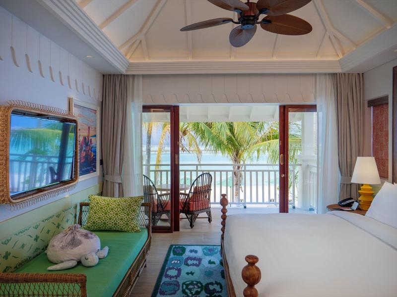 Jasny pokój hotelowy z otwartym balkonem, widokiem na palmy i morze pod białym niebem.