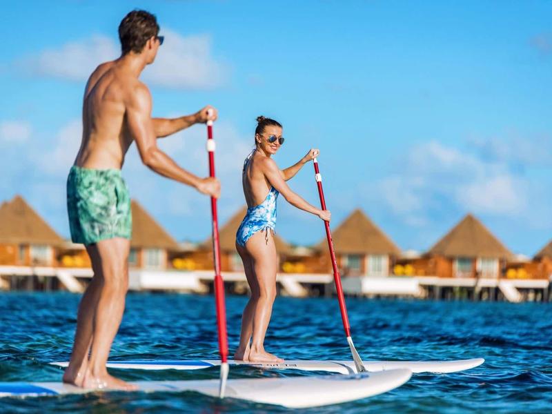Zwei Personen stehen auf Paddleboards im Wasser, Hintergrund mit blauem Himmel und Hütten.