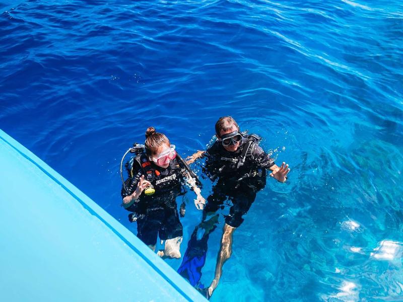 Zwei Taucher im blauen Wasser neben einem Boot, beide tragen Tauchausrüstung.