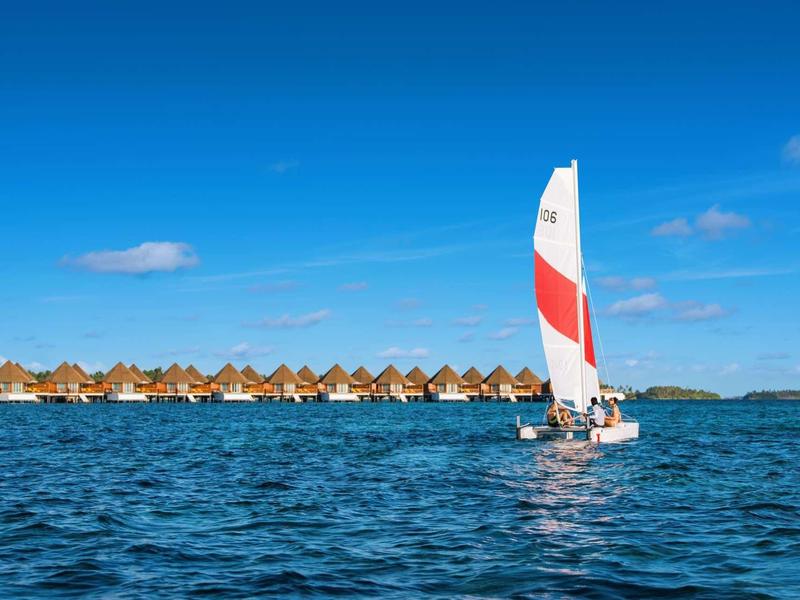 Segelboot mit weißem und rotem Segel auf blauem Wasser vor einer Reihe von Strohdachbungalows.