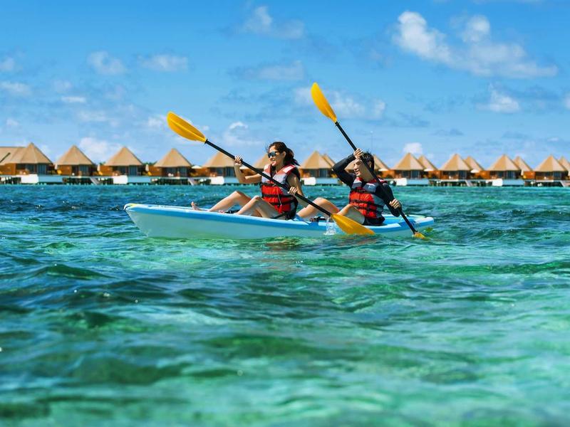 Zwei Personen paddeln im blauen Wasser in einem Doppelkajak bei klarem Himmel und Strandbungalows.