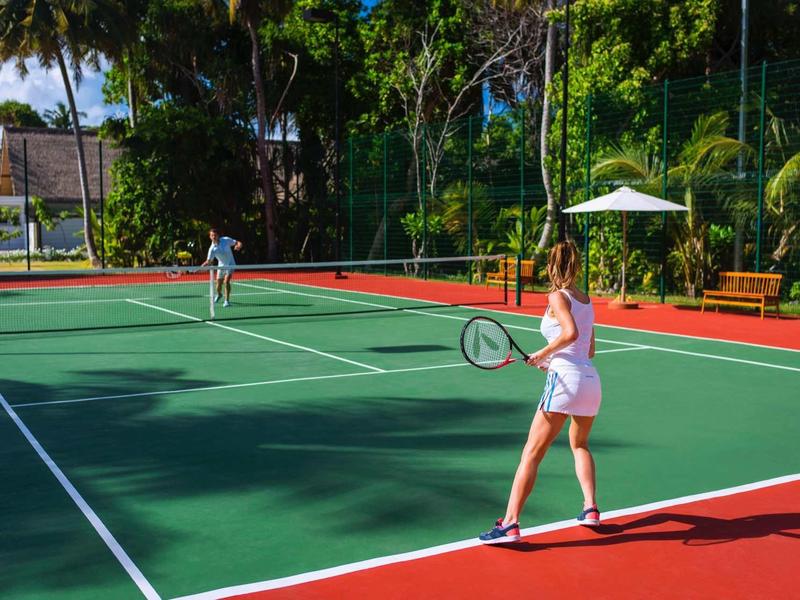 Zwei Personen spielen Tennis auf einem sonnigen Außenplatz mit grüner Spielfläche und rotem Rand.