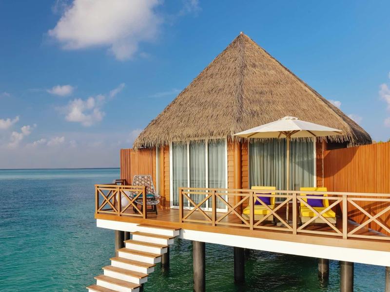 Bungalow mit Strohdach auf Stelzen über klarem blauem Meer, Terrasse mit Sonnenschirm und Treppe ins Wasser.