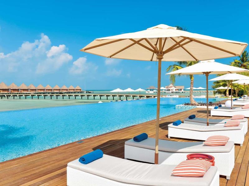 Pool mit Holzdeck, weißen Liegen, Sonnenschirmen und blauem Himmel im Hintergrund.