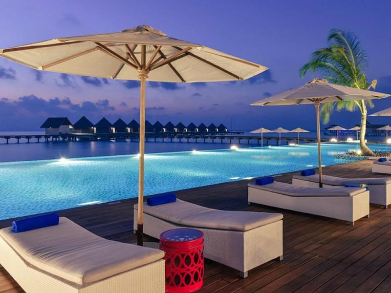 Luxuriöser Poolbereich mit Liegen, Sonnenschirmen und Blick auf Wasserbungalows bei Dämmerung.