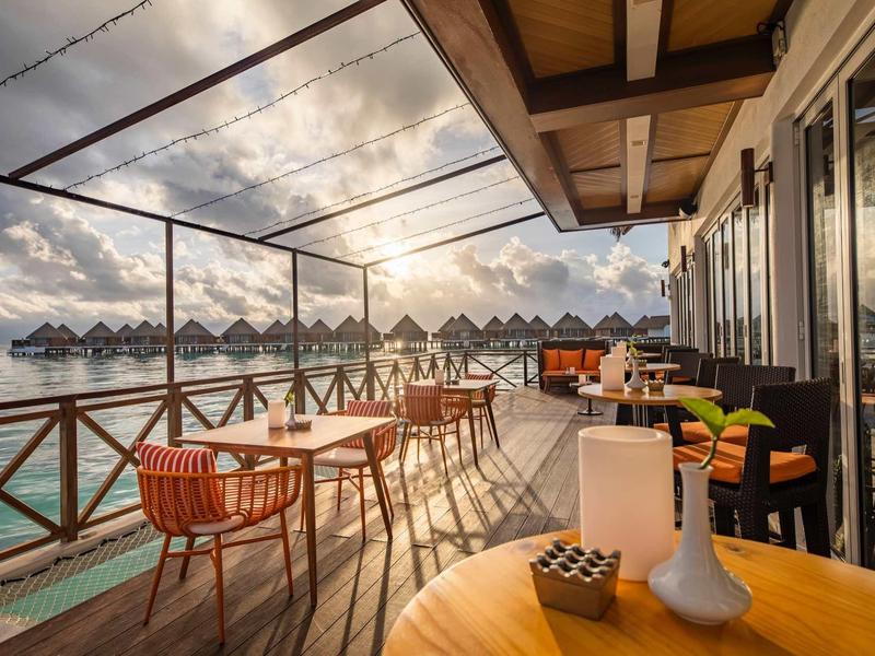 Terrassenbereich mit Holztischen und Stühlen, Blick auf Wasser und überdachte Bungalows im Sonnenlicht.
