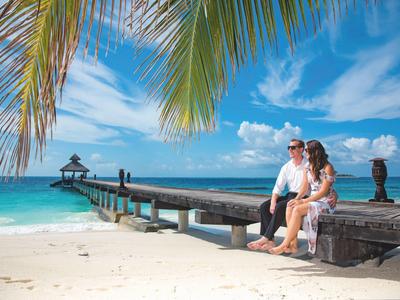 Paar sitzt am Strand auf Steintisch, blauer Himmel, Palmenblätter und langer Steg ins Meer.