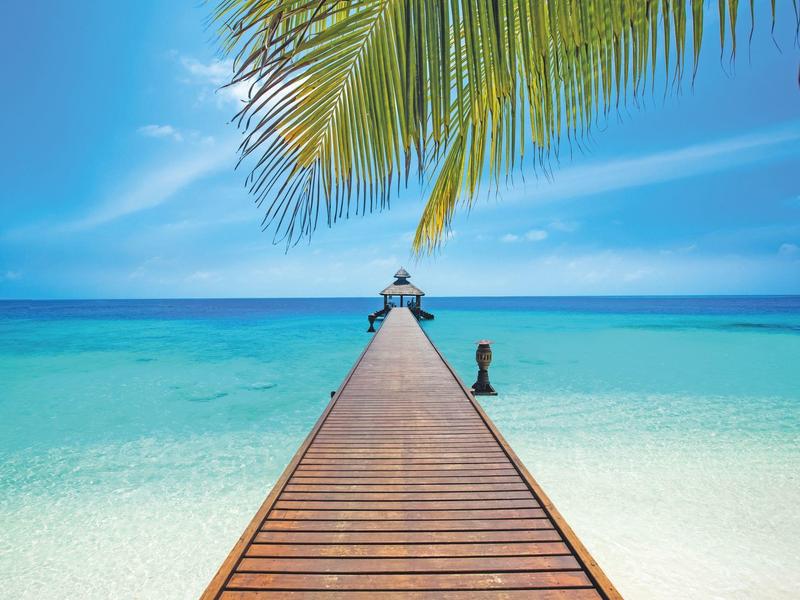 Un molo di legno si estende su sabbia bianca e mare turchese sotto un cielo azzurro.