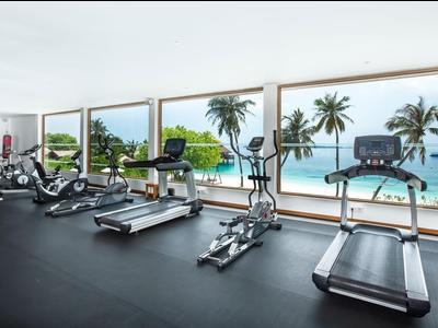 Fitnessraum mit Trainingsgeräten mit Blick auf das Meer und Palmen durch große Fenster.