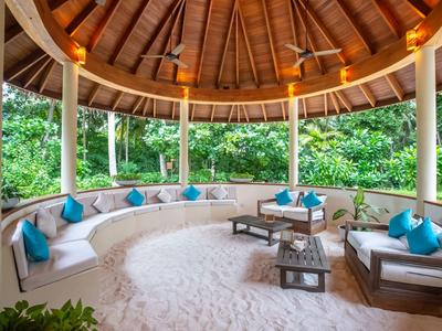 Open lounge met zandvloer en houten dak, omgeven door groene vegetatie.