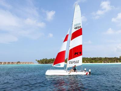 Segelboot mit rot-weißen Segeln auf blauem Meer vor tropischer Insel.