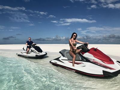 Zwei Personen auf Jetskis im klaren, flachen Wasser unter blauem Himmel.