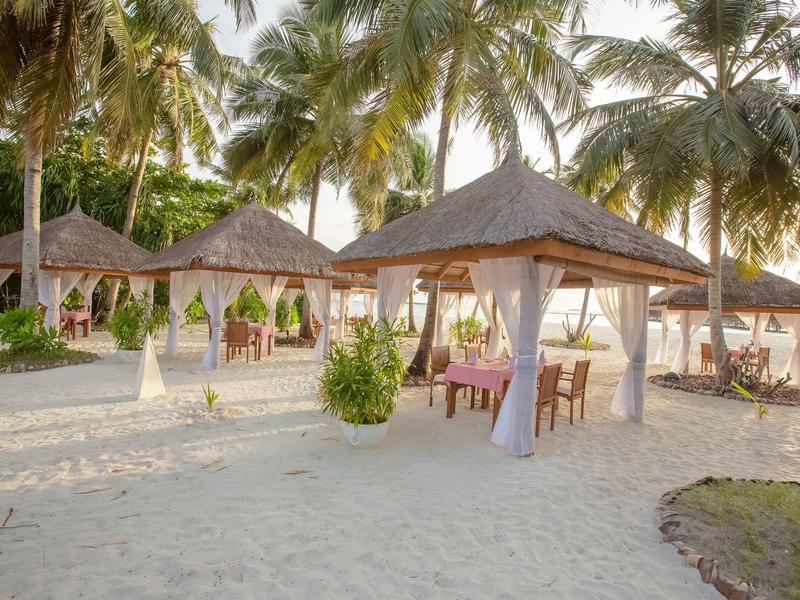 Strandrestaurant met rieten dak en tafels tussen palmboom op wit zand.