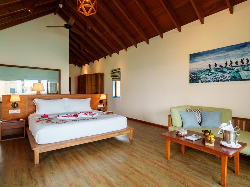 Lichte hotelkamer met houten vloer, groot bed en zithoek met tafel en drankjes.