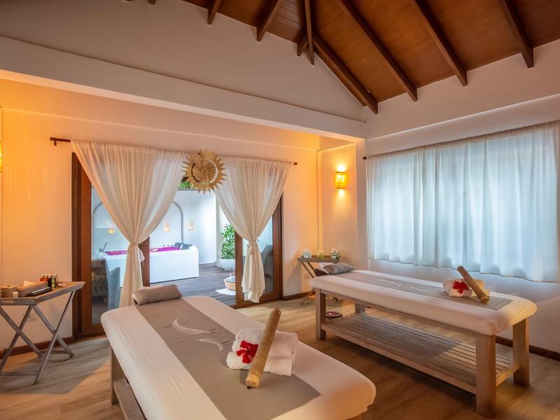 Lichte spa-kamer met twee massagetafels, witte gordijnen en warme houtachtige sfeer.