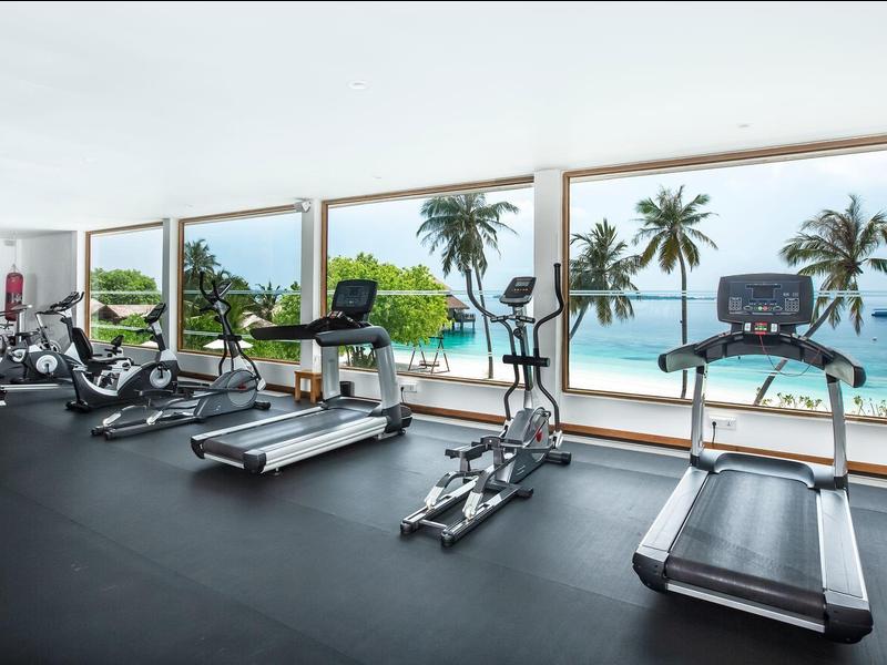 Fitnessruimte met moderne apparatuur en panoramische ramen met uitzicht op strand en palmbomen.