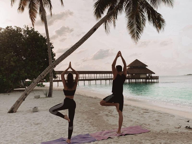 Twee mensen doen yoga op het strand met palmbomen en een pier op de achtergrond.