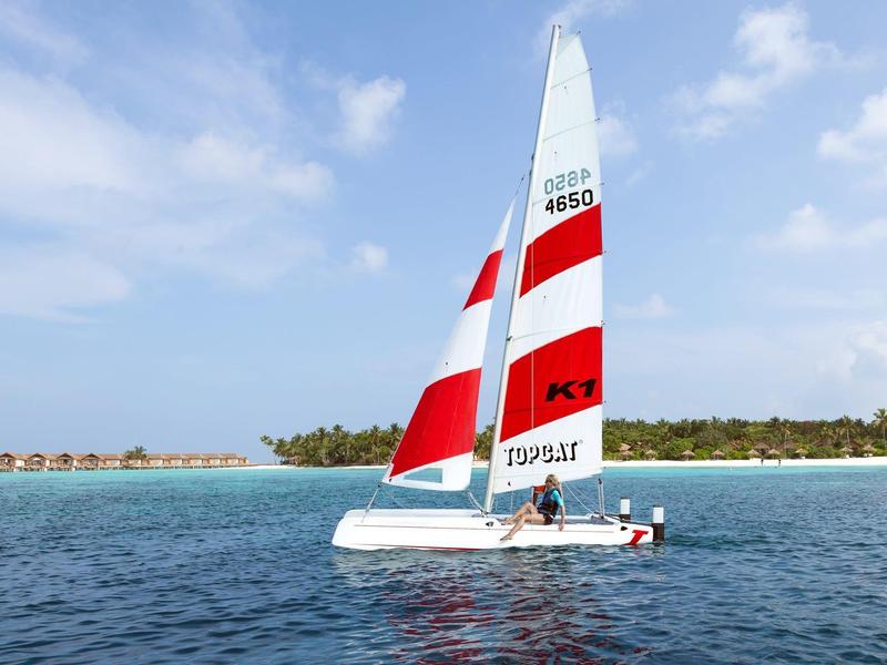 Zeilboot met rood-witte zeilen op blauwe zee bij een eiland met palmbomen.