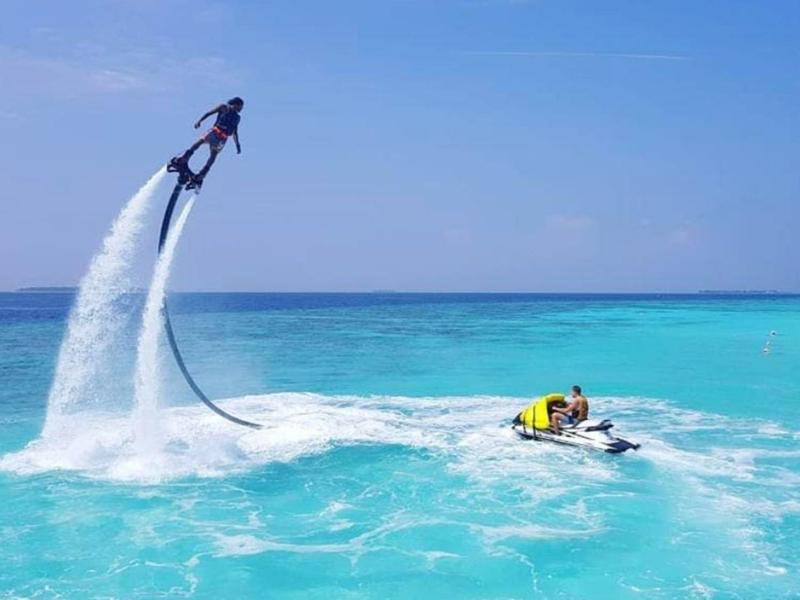 Persoon vliegt met flyboard boven turquoise zee naast jetski onder blauwe lucht.