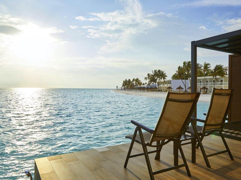 Zwei Liegestühle auf einer Terrasse mit Blick auf das Meer und Palmen bei Sonnenuntergang.