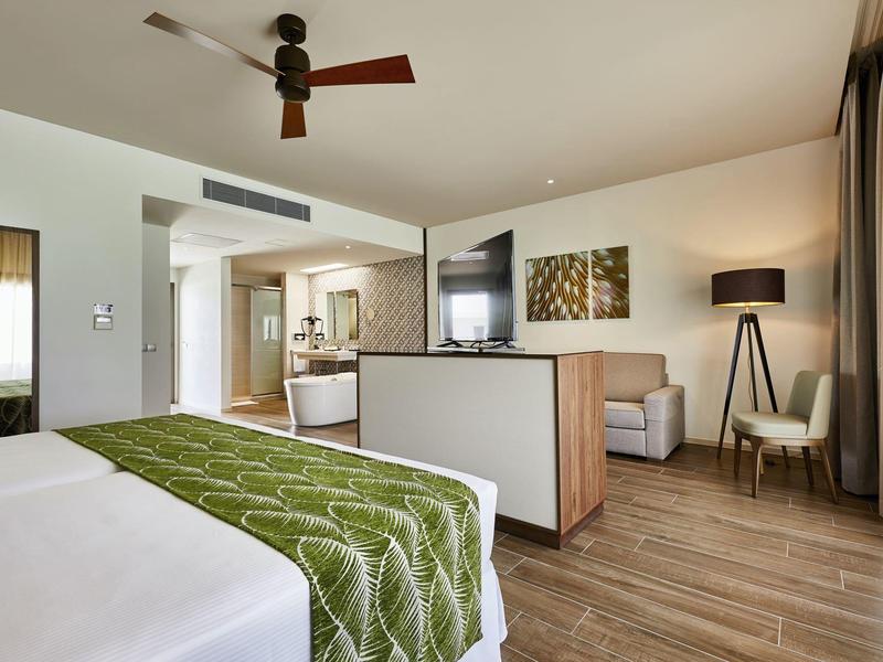 Helles Hotelzimmer mit Doppelbett, Wohnbereich, Fernseher und großem Fenster mit Vorhängen