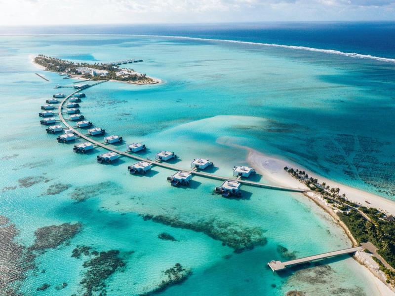 Überwasserbungalows vor einer Insel mit klarem türkisfarbenem Wasser und weißem Sandstrand.